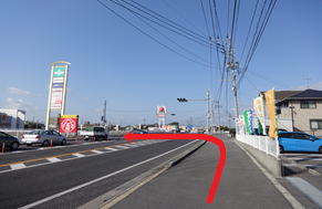 電柱や住宅が並ぶ歩道を直進し、前方の信号がある交差点を左に曲がって片側一車線の車道へと入る経路を、路面上に大きく描かれた赤い矢印で強調した、商業施設の看板やのぼり旗が見える晴天の日の風景写真