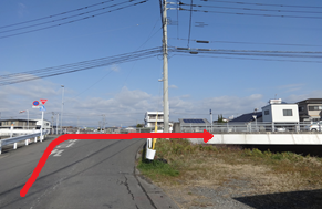 青空の下、白いガードレールが設置された緩やかな坂道を上り、その先にある橋を渡って右方向へと進む経路を、路面上に大きく描かれた太い赤色の矢印で強調して示した写真