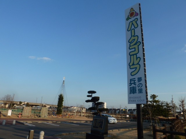 夕方の澄んだ青空の下に、「パークゴルフ 夢の里 兵庫」と縦書きで大きく記された白い看板が立っており、背景には駐車場や松の木、遠くの建物や高架橋が広がる、施設の入口付近の様子の写真