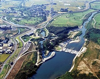広大な平野を流れる大きな河川と、その周辺に整備された水辺の公園や遊歩道、住宅地、田園風景を高い位置から俯瞰したもので、自然と人工物が調和した緑豊かな景観が広がる空撮写真