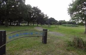 緑豊かな芝生の広場が広がる公園の入口付近で、木製の支柱に支えられた緩やかな曲線を描く透明な案内板が手前に設置され、奥には木立や散策路が点在している写真