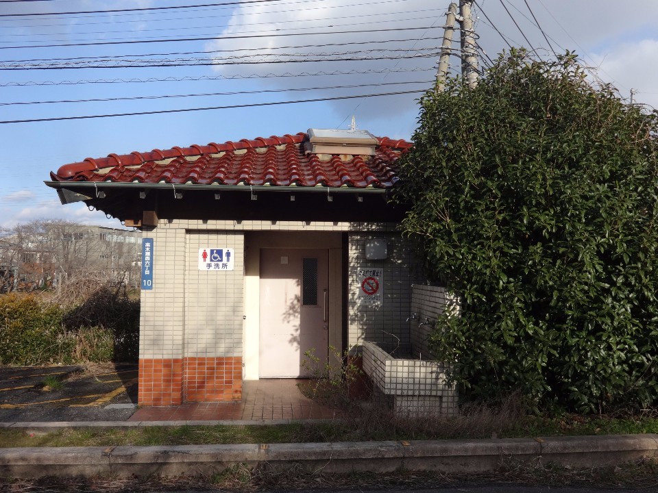 青い空の下、赤茶色の瓦屋根とベージュのタイル壁が特徴的な公衆トイレが建っており、入り口横には手洗い場と大きな常緑樹が配置され、建物の壁面には多機能トイレを示す案内板が設置されている様子を捉えた写真