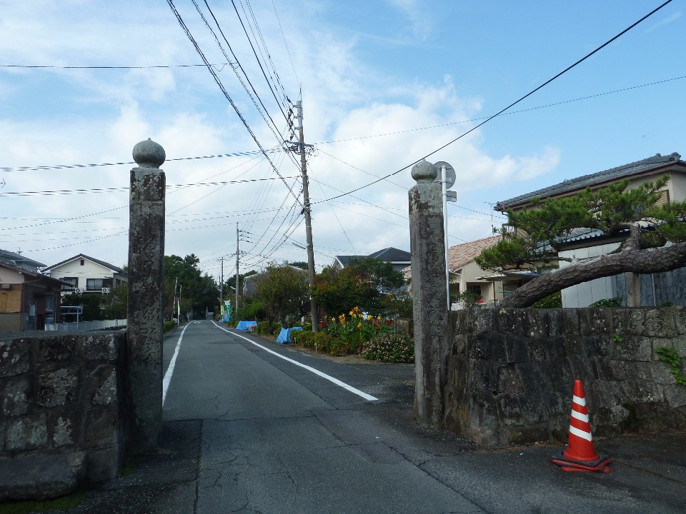 薄曇りの空の下、住宅街を通る細い一本道の入口に、文字が刻まれた古い二本の石柱が門のように立ち、右側の石垣からは立派な松の枝が道へとせり出している写真