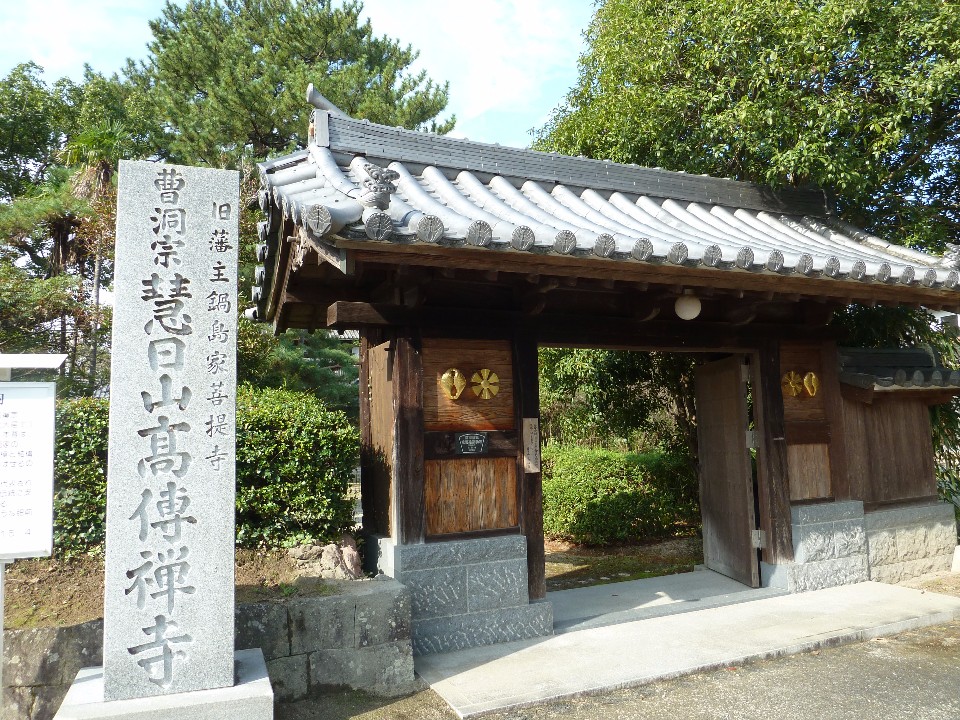 澄み渡る青空と豊かな緑を背景に、旧佐賀藩主鍋島家の菩提寺である「高伝寺」の重厚な瓦屋根の門と、その傍らに「曹洞宗 慧日山 高傳禪寺」と力強く刻まれた大きな石碑が並び立つ、寺院入口の風景を捉えた写真