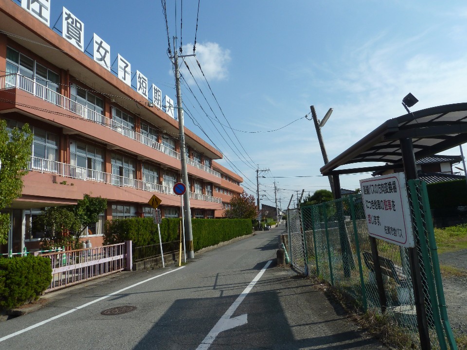 澄み渡る青空の下、左側に「佐賀女子短期大学」と書かれた看板を冠する赤茶色の大きな校舎が建ち、その脇を通る細い一本道の右側にバス停の待合所が設置された、学校周辺の日常的な風景を捉えた写真