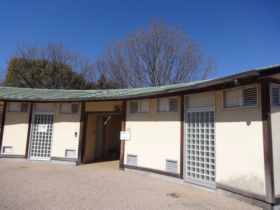 青空を背景に、緩やかにカーブしたクリーム色の外壁と薄緑色の屋根を持つ平屋建ての公衆トイレが建っており、ガラスブロックの窓や入り口付近の貼り紙、背後に並ぶ落葉した木々が特徴的な公園トイレを写した写真