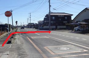 澄み渡る青空の下、住宅が立ち並ぶ片側一車線の直線道路において、左側の歩道から車道を横切り、右前方にある黒い外壁の住宅の前へと進むルートを、赤い矢印で分かりやすく示した道案内の写真