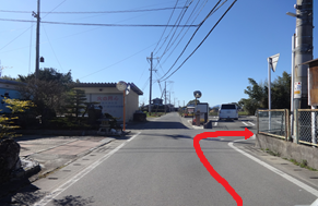 澄み渡る青空の下、民家や倉庫が点在するのどかな一本道において、前方の交差点を右へと曲がり、白い乗用車が停まっている脇の道へと進むルートを、赤い矢印で分かりやすく示した道案内の写真