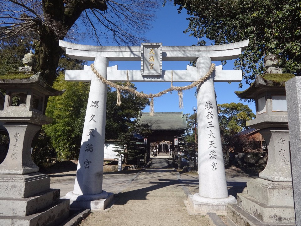 澄み渡る青空の下、注連縄が飾られた白く立派な石造りの明神鳥居が立ち、左右に「常夜燈」と刻まれた石灯籠が並び、その奥へと続く参道の先に瓦屋根の本殿や豊かな緑が佇む、「蛎久天満宮」の静かで厳かな風景を捉えた写真