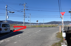 澄み渡る青空の下、遠くに山々を望む開放的な丁字路の交差点において、前方の田園地帯へと続く道を左へと曲がるルートを、赤い矢印で分かりやすく示した道案内の写真