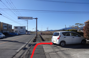 澄み渡る青空の下、遠くに山々を望む直線道路において、頭上の案内標識に従って白い軽自動車が停まっている右側の脇道へと入るルートを、赤い矢印で分かりやすく示した道案内の写真