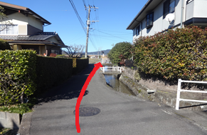 澄み渡る青空の下、住宅の生垣に挟まれた狭い生活道路において、右側に流れる小さな水路に沿って直進し、前方の小さな白い橋の脇を通り抜けるルートを、赤い矢印で分かりやすく示した道案内の写真