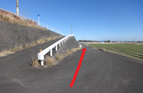 澄み渡る青空の下、左側に堤防へと続くガードレール付きの坂道があり、その脇の平坦な舗装路を田園地帯に向かって真っ直ぐ進むルートを、赤い矢印で分かりやすく示した道案内の写真