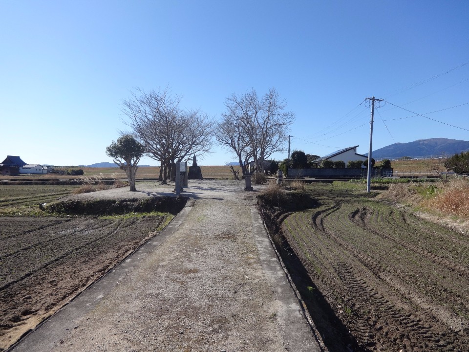澄み渡る青空の下、両側に耕作された田畑が広がる未舗装の道を進んだ先に、数本の冬枯れの木々に囲まれた小さな石碑や案内板が立つ、広々とした御館の森の写真