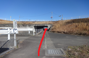 澄み渡る青空の下、堤防の道路の下をくぐり抜けるコンクリート造りの狭いトンネル（アンダーパス）へと続く道を、赤い矢印で真っ直ぐ進むように分かりやすく示した道案内の写真
