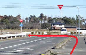 澄み渡る青空の下、丁字路の交差点において、一時停止の標識がある右側の道から合流し、前方の山々や住宅が見える左方向の道へと進むルートを、赤い矢印で分かりやすく示した道案内の写真