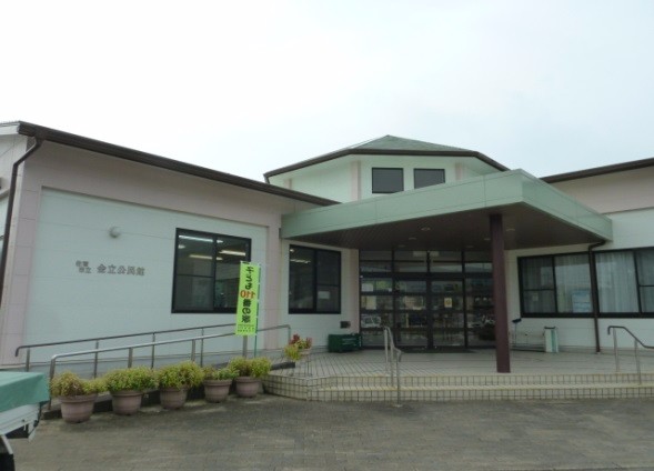 曇り空の下で「佐賀市立金立公民館」と壁面に書かれた平屋建ての白い建物があり、スロープや鉢植えが並ぶ入り口付近には緑色ののぼり旗が立てられ、多角形の屋根構造が特徴的なエントランス周辺を捉えた外観写真