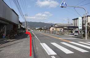 晴天の下で山並みを背景に直線的な道路が続き、手前の横断歩道から左側の細い路地へと進むルートが赤い矢印で示されており、沿道には住宅や「和泉公民館」の看板が並ぶ風景を案内する、道順を説明した写真