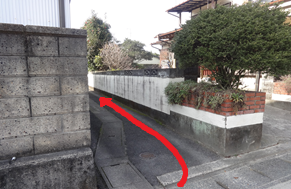 住宅街にあるブロック塀と白い壁に挟まれた非常に細い路地の入り口を捉えており、手前から奥に向かって進むべきルートが赤い曲線状の矢印で強調して描かれた、目的地までの道順を詳細に案内する写真