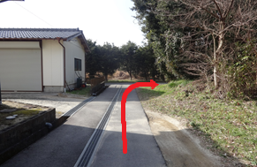 左側にシャッター付きの白い建物があり、舗装された細い道が奥へと続き、突き当たりを右方向に進むよう赤い矢印で示された、木々に囲まれた静かな場所にある目的地のルートを案内する写真