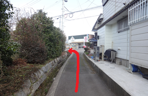 左側に石垣と生け垣、右側に白い住宅の壁がある舗装された細い路地を進み、前方の突き当たりにある建物が並ぶ広い通りへ出て左折するよう赤い矢印で道順が示された、住宅街の歩行ルートを案内する写真