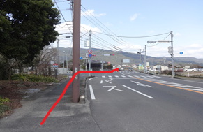 左側に木々が茂る歩道があり、前方の交差点で「バス」と路面に書かれた広い車道へ合流して右方向へ進むルートが赤い矢印で示されており、遠くに山並みが広がる晴天の下の道路風景を捉えた道順案内写真