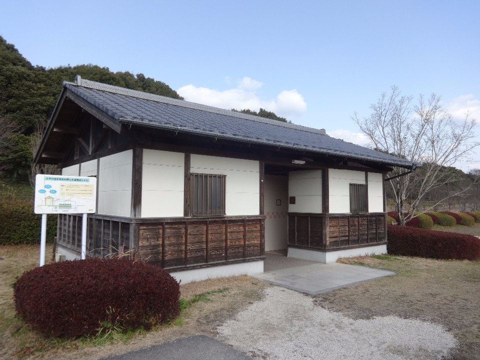 青空の下、白い壁と木の質感を活かした腰壁が特徴的な瓦屋根の平屋建て公衆トイレが建っており、左手には説明看板、周囲には低木や芝生が整備された外観写真
