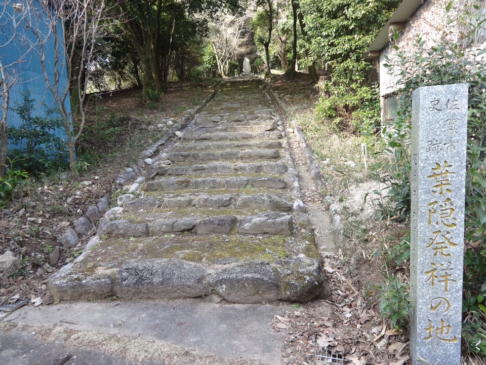 木々に囲まれた静かな場所に、苔が生した古い石段が真っ直ぐ上方へと続き、右手前には「史跡 佐賀市 葉隠発祥の地」と金色で刻まれた立派な石碑が佇む、厳かな雰囲気の風景写真