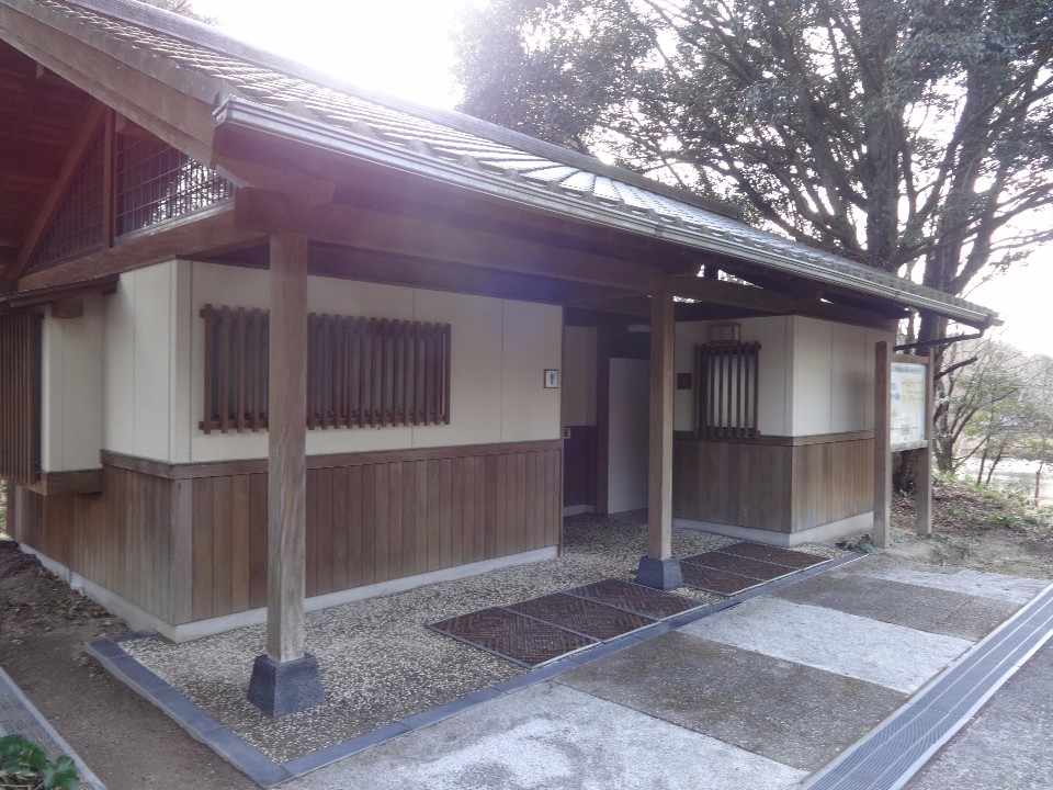 木々に囲まれた自然豊かな場所に建つ、伝統的な瓦屋根と木の質感、白い壁が調和した平屋建ての公衆トイレで、入口には屋根を支える太い木柱が並び、手前には案内看板が設置された外観写真