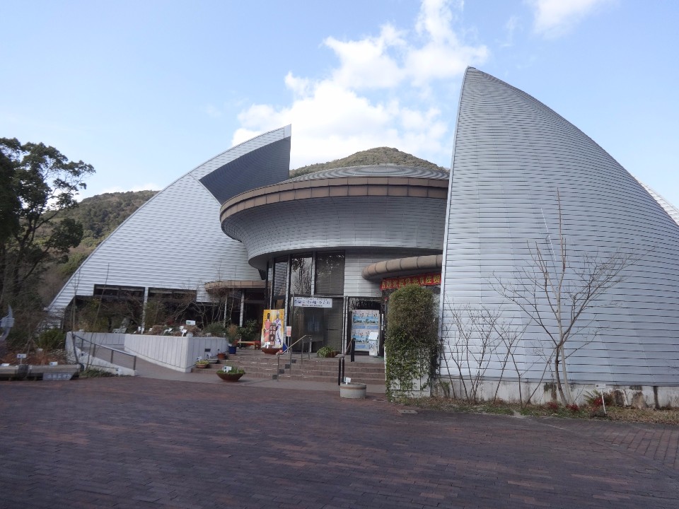 背後にそびえる山並みを背景に、左右に広がる巨大な三角形の尖った外壁と中央の円形構造が組み合わさった近未来的で個性的なデザインの建物が建ち、広場からスロープや階段が続くエントランス周辺を捉えた徐福長寿館の外観写真
