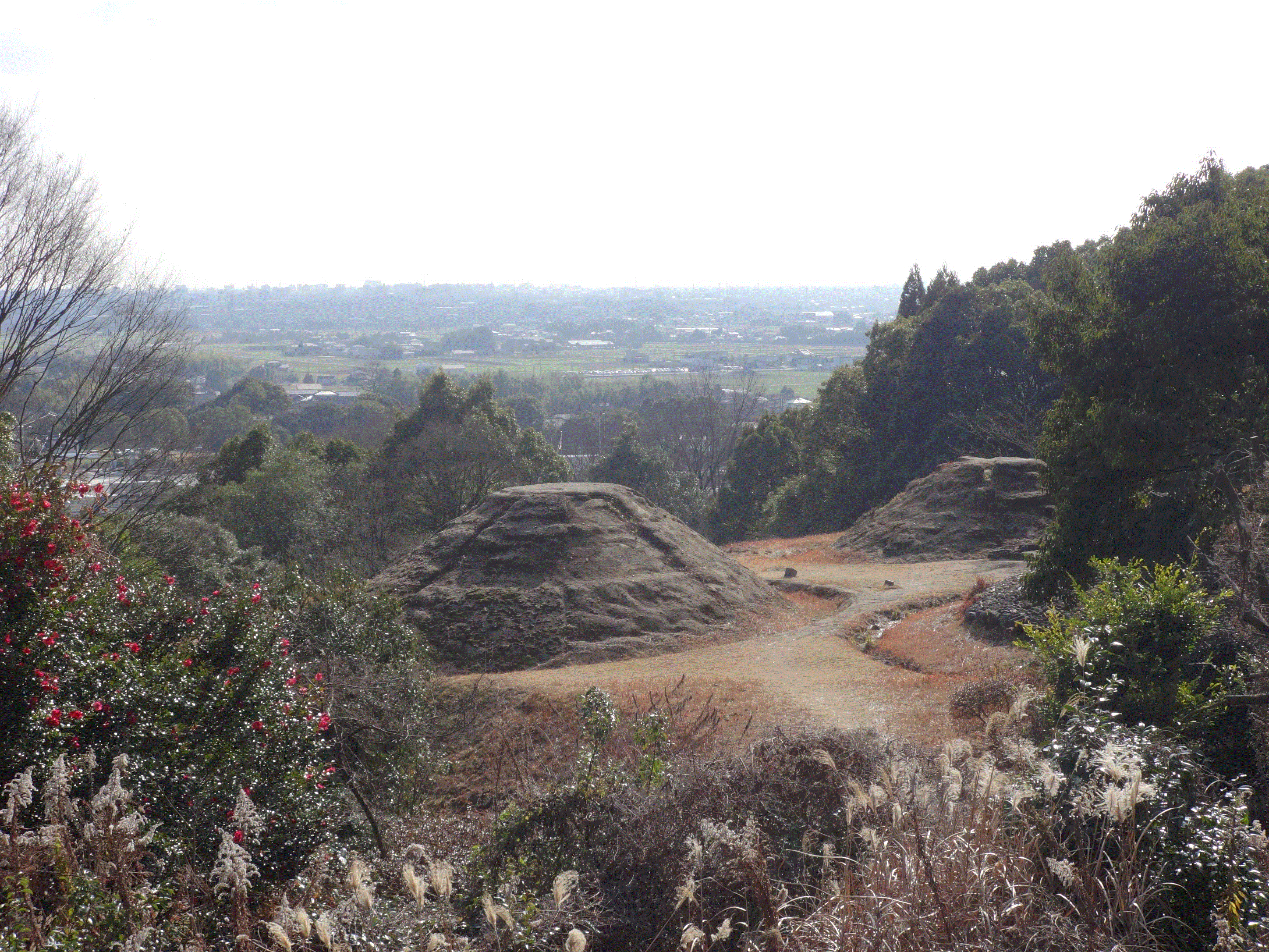 高台から見下ろす視点で、手前の木々の間に円錐台のような形をした大小二つの土盛りが並び、その奥には広大な佐賀平野の田園風景と街並みが遠くまで広がる、歴史的な金立銚子塚古墳の形状を捉えた風景写真