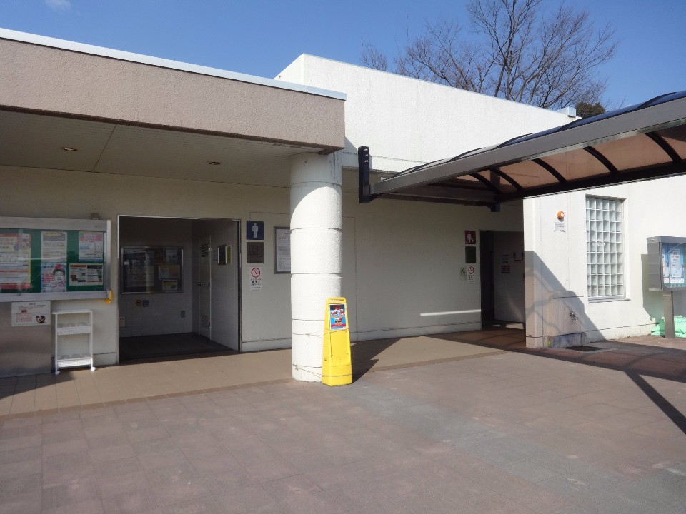 明るい日差しが差し込む白い外壁の平屋建て公衆トイレがあり、左側の入口付近には掲示板、中央の太い円柱横には黄色の清掃中看板が置かれ、右側にはアーチ型の屋根が続くトイレの屋外写真