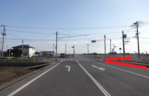 晴天の下、二車線の真っ直ぐな道路が地平線まで続き、前方の交差点を右折するルートが太い赤い矢印で示されており、周囲には田畑や住宅が点在する開放的な風景の中で次の進行方向を案内する道順案内写真