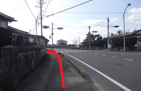 左側に石垣と古い民家の塀が続く歩道があり、前方の信号機がある交差点を左折するルートが赤い矢印で示されており、遠くに住宅が点在する晴天の下の静かな街並みを捉えた、目的地への道順を案内する写真