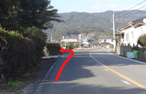 晴天の下、遠くに山々を望む佐賀市の二車線道路において、左側の茂みの手前にある「宮分」と書かれた看板の地点で、路面に描かれた赤い矢印に従って左の細い脇道へと曲がる様子を示した道順説明の写真