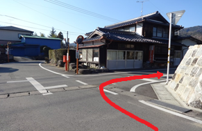歴史的な木造建築が建つ街角の十字路において、角に設置された赤い丸型ポストとカーブミラーを目印に、路面に描かれた赤い矢印の指示に従って右方向へと曲がる、佐賀市のウォーキングコースの道順を説明するための写真