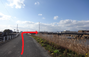 青空の下、周囲には枯れたすすきや遠くの建物が見える、川沿いの舗装された平坦な道を直進した後に右折して橋を渡るルートを、写真上に描かれた太い赤い矢印で示している写真