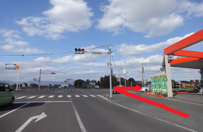 青空が広がる見通しの良い交差点で、右側のガソリンスタンド側の歩道から車道へ向けて右折し、横断歩道のある幹線道路へと向かうルートを太い赤い矢印で示した写真