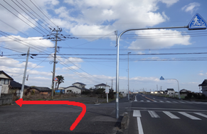 青空の下、住宅地が広がる見通しの良い道路を直進し、横断歩道の手前にある細い路地を左へ曲がるルートを、路面上に描かれた太い赤い矢印で示した写真