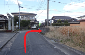 周辺の民家や電柱が立ち並ぶ景色を背景に、住宅街を通る細いアスファルトの道を直進し、左側に鳥居が見える丁字路を右に曲がるルートを、路面上に描かれた太い赤い矢印で示した写真