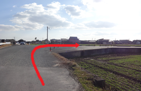 周囲に民家や畑が点在するのどかな風景を背景に、田園地帯の舗装路を直進し、右側に広がる田畑へ向かって右折するルートを、路面上に描かれた太い赤い矢印で示した写真