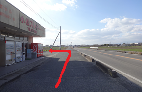 幹線道路沿いの歩道を直進し、自動販売機が並ぶ「小松ストアー」という看板の店舗の先の路地を左に曲がるルートを、アスファルトの上に太い赤い矢印で示した写真