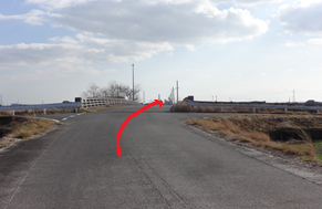 晴れ渡った空の下、周囲に広がる冬枯れの草地や遠くの電柱を背景に、見通しの良いアスファルトの道をまっすぐ進み、前方に現れる小さな橋を渡るルートを、路面上に描かれた太い赤い矢印で示した写真