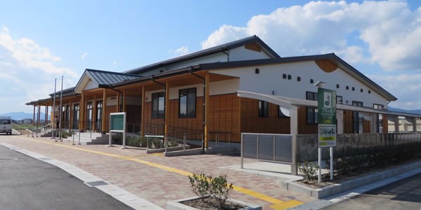 青い空と白い雲の下に建つ、白い壁と温かみのある木材の外壁が特徴的な平屋建ての建物と、その手前に整備された点字ブロックのある歩道や看板などが写し出された、新栄公民館の写真