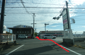 晴れ渡る空の下、道路の向かい側に店舗や看板が並ぶ交差点付近の風景を捉えており、手前の白線から交差点を渡って奥の細い道へと進む経路を赤い矢印で示している写真