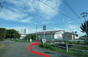 青空の下、左側に緑豊かな木々、右側に小さな橋と建物が見える三差路の風景を捉えており、手前の道から右方向へ曲がり、橋を渡って建物の方へと進む経路を赤い矢印で示している写真