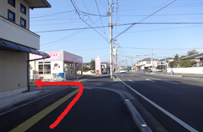 抜けるような青空の下、片側一車線の広い道路沿いにある住宅街の風景を捉えており、手前の歩道から左側にある建物の角を曲がる経路を太い赤い矢印で示している写真