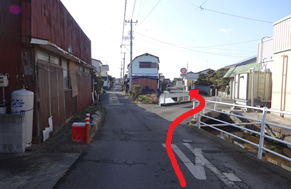 晴れ渡る空の下、左側に年季の入った茶色の建物、右側に白い柵のある水路が続く細い路地の風景を捉えており、手前から右前方にある小さな橋を渡って奥の住宅街へと進む経路を、太い赤い矢印で示している写真