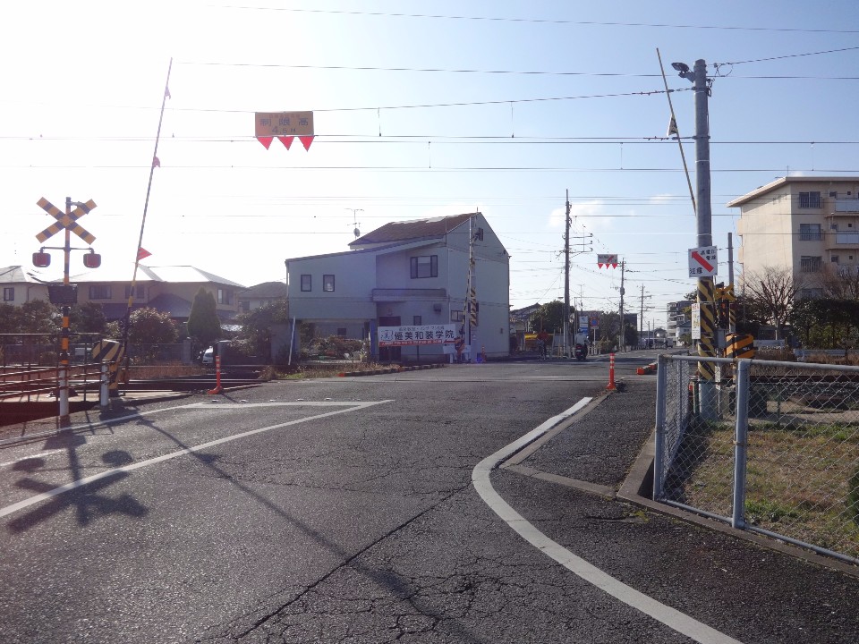 晴天の下、遮断機が上がった状態の線路が横切る踏切の風景を捉えており、手前の道路から踏切を渡った先には一般住宅が立ち並び、遠くまで続く平坦な道の様子や周辺の静かな住宅街の雰囲気を伝えている写真