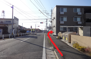 澄み渡る青空の下、歩道の右側に建つ茶色の集合住宅の前を通り、その先の角を右に曲がって建物の裏手へと進む経路を太い赤い矢印で示している写真